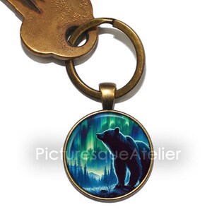 Könnte beinhalten: Ein Schlüsselanhänger aus Bronze mit einem runden Glasanhänger, der eine schwarze Bärensilhouette zeigt, die in einem Wald mit grünem und blauem Nordlicht im Hintergrund steht.