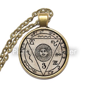 Könnte beinhalten: Eine goldene Kettenkette mit einem runden Anhänger, der ein Schwarzweißbild einer Sonne mit einem Gesicht in einem Dreieck zeigt. Der Anhänger ist von einem Kreis mit schwarzem Text und Symbolen umgeben.