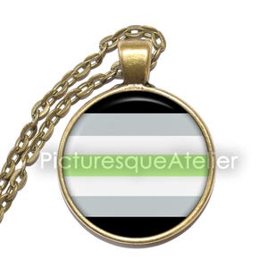 Op de afbeelding: Een ketting met een ronde hanger en een goudkleurige ketting. De hanger toont de Agender pride vlag, met zwarte, grijze, witte en groene horizontale strepen. De tekst "Picturesque Atelier" is op de hanger gedrukt.
