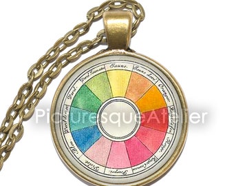 May include: A gold necklace with a round pendant featuring a vintage color wheel with French text. The color wheel is divided into 12 sections, each with a different color and French name. The colors are red, orange, yellow, green, blue, violet, and their variations.