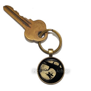 May include: A brass key chain with a round, glass charm featuring a silhouette of a witch flying on a broomstick in front of a full moon. The charm is attached to a brass key ring.