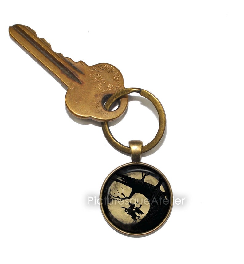 May include: A brass key chain with a round, glass charm featuring a silhouette of a witch flying on a broomstick in front of a full moon. The charm is attached to a brass key ring.