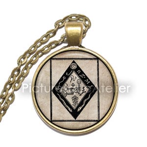 May include: A gold chain necklace with a round pendant featuring a black and white image of a diamond shape with a symbol inside. The symbol includes a hand holding a torch, a snake, a flower, and the text "n.z.m."