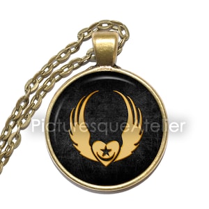 Può includere: Una collana con un ciondolo dorato su sfondo nero che presenta un cuore dorato con una stella al suo interno e ali su entrambi i lati. Il ciondolo è attaccato a una catena dorata.