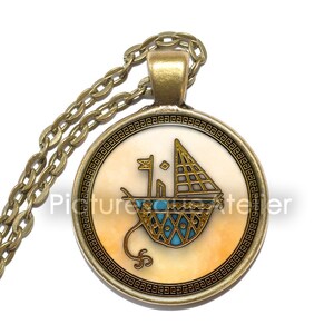 May include: A gold necklace with a round pendant featuring a stylised image of a ship with a sail, surrounded by a decorative border. The pendant is on a cream background with a blue and gold design.