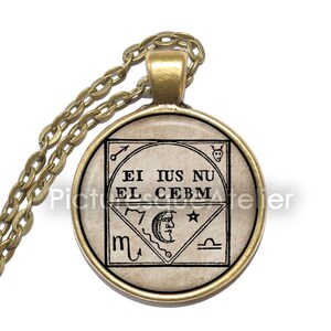 May include: A gold necklace with a round pendant featuring a vintage-style illustration of a symbol with the text "EI IUS NU EL CEBM" and astrological symbols.