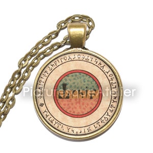 Op de afbeelding: Een gouden ketting met een ronde hanger met een cirkelvormig ontwerp met een rood, oranje en groen patroon erin. De hanger is omgeven door een cirkel met tekst in een oude taal.