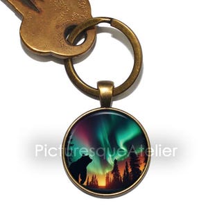 May include: A bronze key chain with a round glass charm featuring a silhouette of a bear standing in front of a colourful aurora borealis display. The aurora is green, pink, and purple with a dark forest in the background.