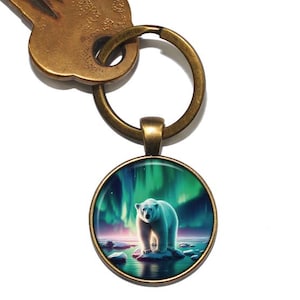 Peut inclure: Un porte-clés avec un charme rond en verre représentant un ours polaire debout sur un lac gelé sous une aurore boréale verte et bleue.