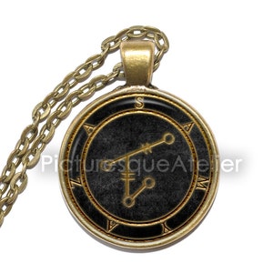 May include: A gold chain necklace with a round pendant featuring a black background and a gold sigil. The sigil is a stylised symbol with a cross and two circles. The pendant is surrounded by a gold circle with the letters "S", "A", "M", and "V" inscribed around it.