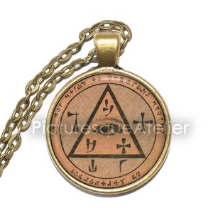 Op de afbeelding: Een gouden ketting met een ronde hanger met een bruin en gouden zegel met een driehoek, een oog en symbolen. De hanger heeft een gouden rand en de tekst " + L Q R A L U N X H T A O R I N H T R W M " rond de rand.