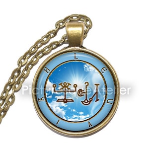 Puede incluir: Un collar dorado con un colgante redondo que presenta un cielo azul con nubes blancas y un sol brillando a trav&eacute;s. El colgante tambi&eacute;n tiene un sello marr&oacute;n con las letras "LACHUAI" alrededor del borde.