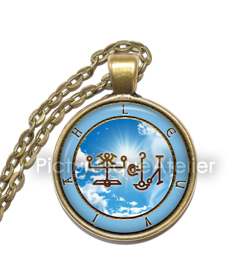 Puede incluir: Un collar dorado con un colgante redondo que presenta un cielo azul con nubes blancas y un sol brillando a trav&eacute;s. El colgante tambi&eacute;n tiene un sello marr&oacute;n con las letras "LACHUAI" alrededor del borde.