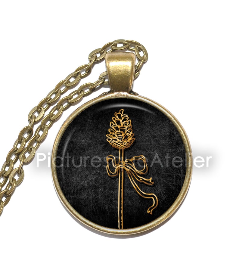 May include: A gold necklace with a round pendant featuring a black background and a gold illustration of a stylized wheat stalk with a bow. The pendant is framed with a gold rim.