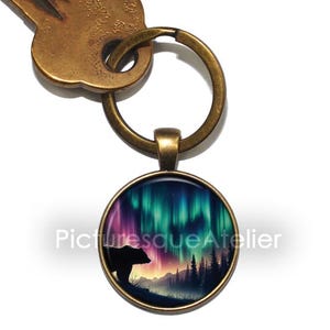 Op de afbeelding: Een bronzen sleutelhanger met een ronde glazen hanger met een silhouet van een beer die voor een kleurrijke aurora borealis-weergave staat. De aurora is een levendige mix van roze, paarse, groene en blauwe tinten. De achtergrond omvat een donker bos en een bergketen.