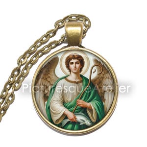 May include: A round pendant necklace featuring an angel in green and white robes, holding a staff and a fish. The gold-coloured pendant hangs from a gold chain. The text "Picturesque Atelier" is visible on the image.