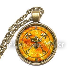 Op de afbeelding: Een gouden ketting met een ronde hanger met een zegel op een vurige oranje achtergrond. De zegel is een gestileerd ontwerp met een cirkel in het midden en twee gekruiste speren met een driehoek aan de bovenkant. De hanger is omgeven door een gouden frame met de letters "B", "A", "L" en "F" rond de rand.