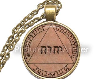 May include: A gold chain necklace with a round pendant featuring a triangle with Hebrew text inside and a circle of Hebrew text around the triangle. The pendant is on a light brown background.