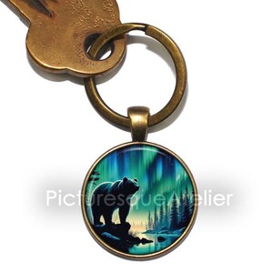Op de afbeelding: Een bronzen sleutelhanger met een ronde glazen hanger met een silhouet van een beer die voor een groen-blauwe aurora borealis staat. De hanger is bevestigd aan een bronzen sleutelring.