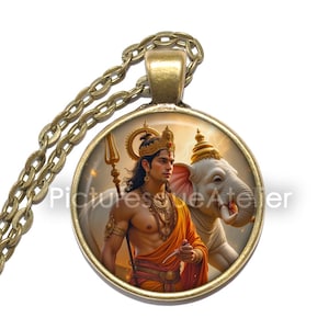 May include: A round pendant necklace with a gold-colored chain. The pendant features a detailed illustration of a deity in orange and gold attire, holding a trident, with a white elephant in the background. The text "Picturesque Atelier" is visible.