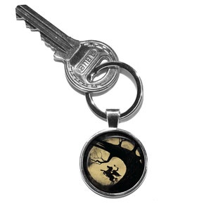 May include: A silver key chain with a round, silver charm featuring a silhouette of a witch riding a broomstick in front of a full moon and a tree.