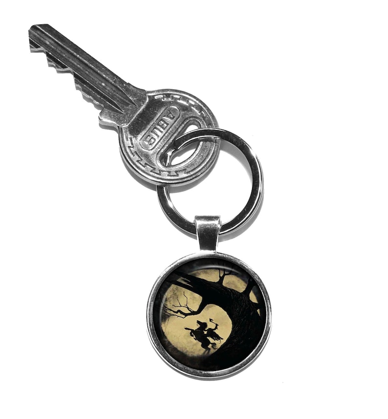 May include: A silver key chain with a round, silver charm featuring a silhouette of a witch riding a broomstick in front of a full moon and a tree.