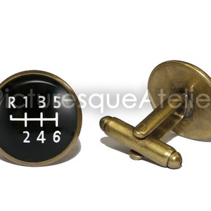 Puede incluir: Un par de gemelos de color dorado con un diseño circular negro que presenta un patrón de cambio de marchas blanco con los números 1, 2, 3, 4, 5, 6 y R.