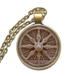 May include: A gold necklace with a round pendant featuring a detailed image of a wooden wheel with intricate carvings. The pendant is surrounded by a gold rim.