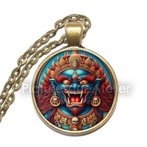 Può includere: Una collana con catena dorata con un ciondolo rotondo in vetro che presenta un'illustrazione colorata di una divinità blu con un'espressione feroce, circondata da fiamme e teschi.
