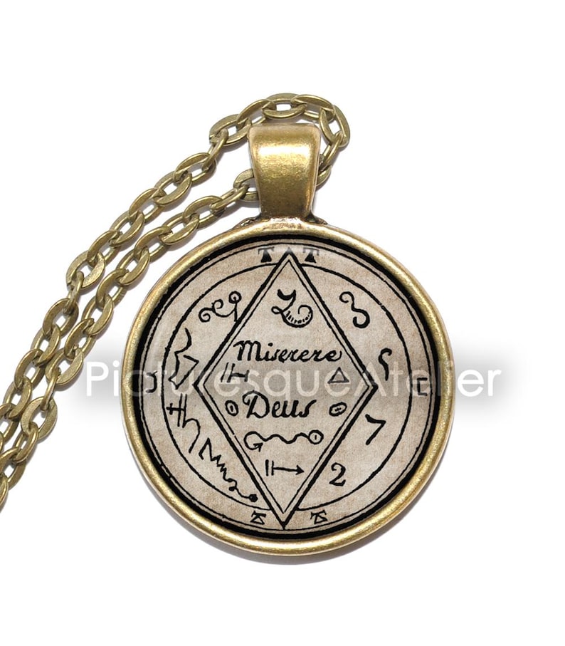 May include: A gold chain necklace with a round pendant featuring a black and white image of a sigil with the text "Misere Deus" and other symbols.