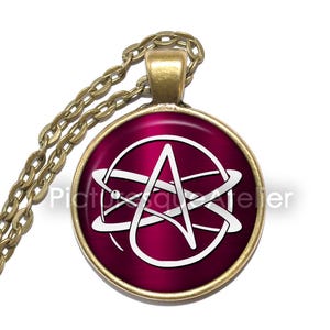 May include: A necklace with a round pendant featuring a white stylized letter A surrounded by two white circles on a dark red background. The pendant is attached to a gold chain.