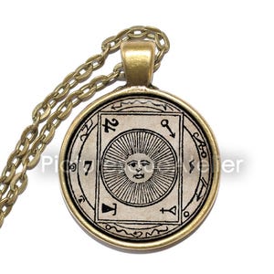 Puede incluir: Un collar de cadena dorada con un colgante redondo que presenta una ilustración en blanco y negro de un sol con cara. El colgante está rodeado por un círculo de texto y símbolos.