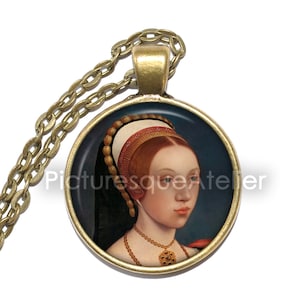 Può includere: Una collana con catena dorata con un ciondolo rotondo che presenta un ritratto di una donna con i capelli rossi che indossa un copricapo bianco e dorato. Il ritratto è su uno sfondo blu scuro.
