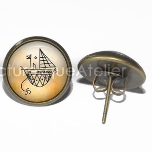 May include: A pair of bronze earrings with a small round glass cabochon featuring a black and white line drawing of a sailboat with the text "IM-U-A-MOU" on a yellow and orange background.