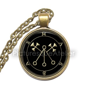 May include: A gold necklace with a black pendant featuring a gold sigil with the letters "M A S B A" around the edge. The sigil depicts a circle with four points, each with a circle at the end and a cross at the top.