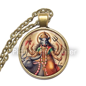 May include: A gold necklace with a round pendant featuring a colorful image of a Hindu goddess with a boar head riding a black bull. The goddess is holding a trident and a staff.