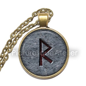 Op de afbeelding: Een gouden ketting met een ronde hanger met een rood runesymbool op een grijze stenen achtergrond.