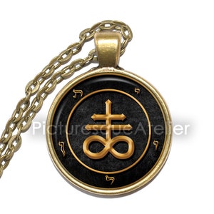 May include: A gold pendant necklace with a black and gold round charm featuring a sigil with a cross and an infinity symbol. The charm is surrounded by Hebrew letters.
