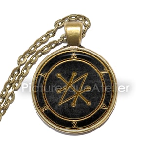 Op de afbeelding: Een gouden ketting met een ronde hanger met een zwarte achtergrond en een gouden zegel. Het zegel is een geometrisch ontwerp met een stervorm en lijnen die naar buiten stralen.