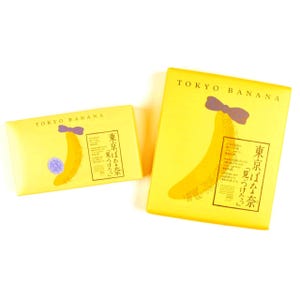 以下が含まれることがあります： TOKYO BANANAと書かれた2つの黄色い長方形の箱。紫色のリボンをつけた黄色いバナナのイラストが描かれています。パッケージには日本語のテキストがあり、小さい方の箱には紫色の小さな円形のシールが貼られており、明るく統一されたデザインです。