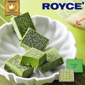 May include: Square pieces of green matcha Nama Chocolate by ROYCE' are arranged in a white scalloped bowl. The chocolate cubes are dusted with green powder, and one piece shows a smooth interior. A box of the chocolate is visible nearby.