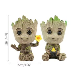May include: Two Groot figurines, each approximately 5 cm tall, are shown. One holds a yellow flower, the other cradles a yellow bird. The figures have a detailed, textured appearance with brown and green tones.