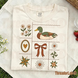 May include: Cream-colored t-shirt featuring a grid design with a duck, daisies, a bow, and other decorative elements. The shirt has a crew neck and the brand name "Comfort Colors" is visible on the tag.