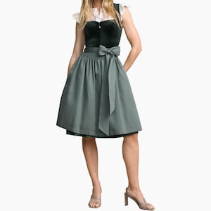 Op de afbeelding: Een jurk met een donkergroen fluwelen lijfje en een witte blouse met korte mouwen en ruches bij de schouders. De jurk heeft een geplooide rok in een gedempte saliegroene kleur, geaccentueerd door een bijpassende brede stoffen ceintuur met een strik.