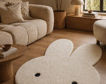 May include: A cream-colored, bunny-shaped rug with black eyes and a black cross for a mouth. The rug is made of a soft, plush material and is placed on a hardwood floor. The rug is a decorative item for a living room.