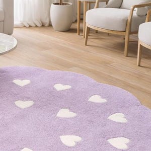 May include: A round, lavender-colored area rug with a scalloped edge, featuring a repeating pattern of small, white, heart-shaped motifs across the entire surface. The rug has a soft, plush texture and is positioned on a light wood floor.