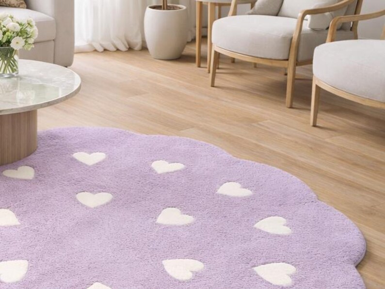 May include: A round, lavender-colored area rug with a scalloped edge, featuring a repeating pattern of small, white, heart-shaped motifs across the entire surface. The rug has a soft, plush texture and is positioned on a light wood floor.