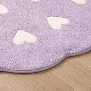 May include: A lavender-colored, heart-shaped rug with white heart accents. The rug has a soft, plush texture and a scalloped edge. The rug is placed on a wooden floor.