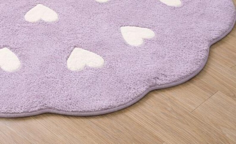 May include: A lavender-colored, heart-shaped rug with white heart accents. The rug has a soft, plush texture and a scalloped edge. The rug is placed on a wooden floor.