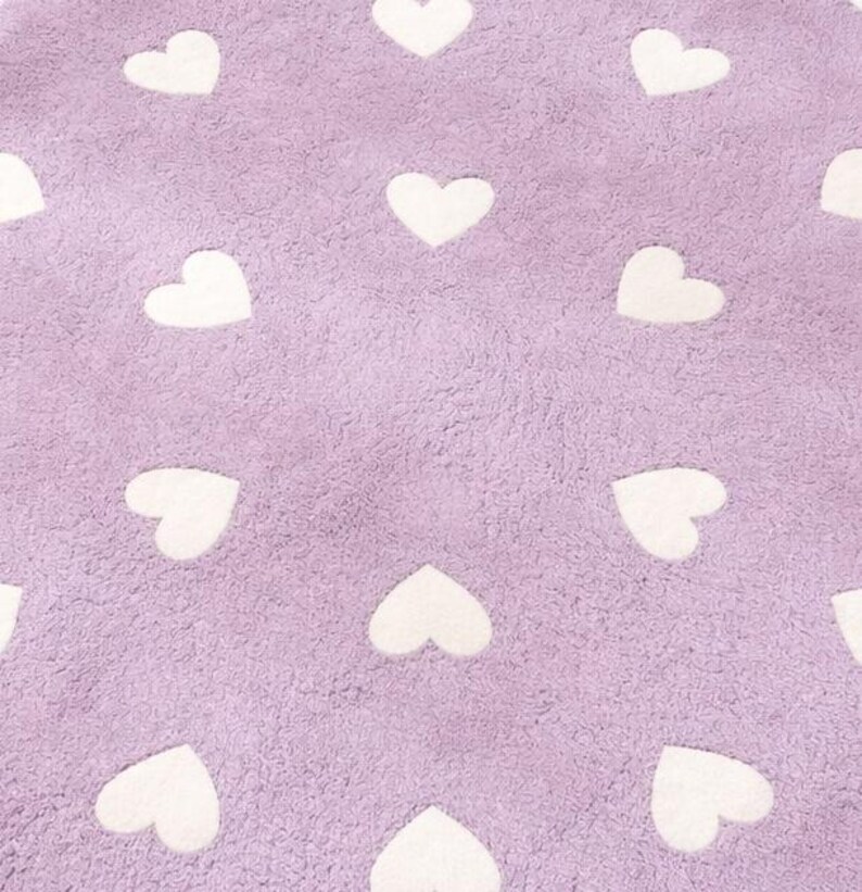 May include: A soft, textured lavender fabric featuring a repeating pattern of white heart shapes arranged in a grid. The material has a plush, fuzzy surface texture throughout, with the white hearts contrasting against the light purple background.
